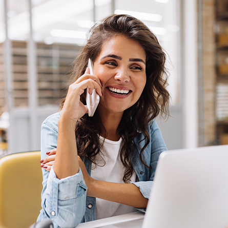 A woman at a desk smiles while talking on her phone.