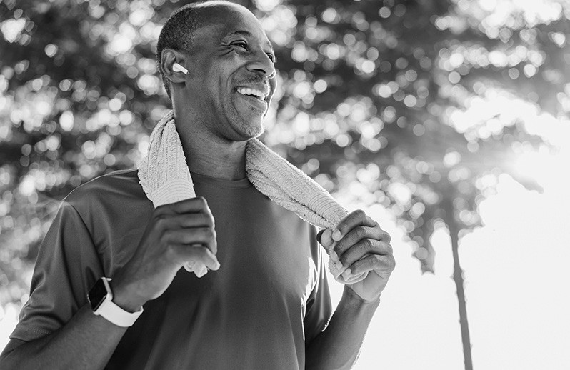 A man walking at the park with towel around his neck and headphone in ears smiling