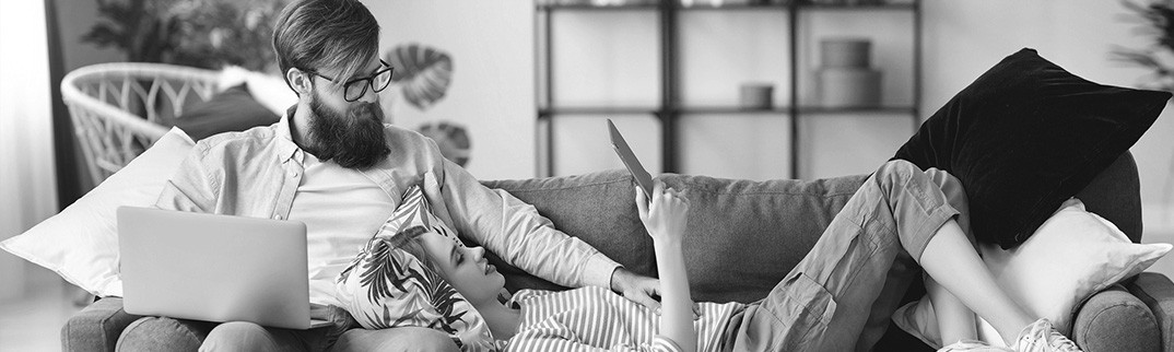 A woman laying on a couch on tablet with man sitting with laptop looking over at her tablet