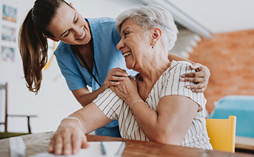 A woman smiles as she hugs another woman who sits at a table.